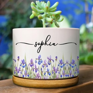 Pode incluir: Um vaso cilíndrico de cerâmica branca com um padrão repetitivo de flores de íris roxas e caules verdes na parte inferior. O nome "sophia" está impresso no centro em letra cursiva preta. O vaso repousa sobre uma base de bambu.