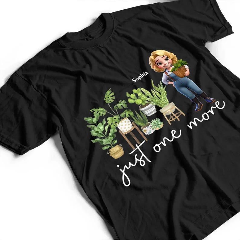 Puede incluir: Camiseta negra con una ilustraci&oacute;n de dibujos animados de una mujer sosteniendo una cesta de plantas, rodeada de varias plantas en macetas. El texto "just one more" est&aacute; escrito en cursiva. El nombre "Sophia" est&aacute; encima de la mujer.
