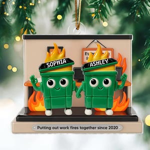 May include: A green and yellow Christmas ornament featuring two cartoon trash cans with flames on their heads. The ornament has the names "Sophia" and "Ashley" on the trash cans. The base reads "Putting out work fires together since 2020."