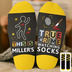 May include: A pair of black socks with yellow toes and heels. The socks feature crime scene graphics, including a chalk outline, footprints, and a bloody knife. Text on the socks reads "THIS IS MILLER'S" and "TRUE CRIME WATCHING SOCKS."
