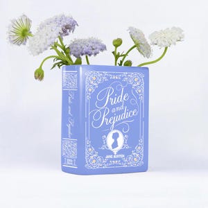 Può includere: Un vaso in ceramica blu chiaro a forma di libro, con il titolo "Pride and Prejudice" e il nome dell'autrice, Jane Austen. Contiene fiori bianchi e viola, creando una composizione floreale decorativa.