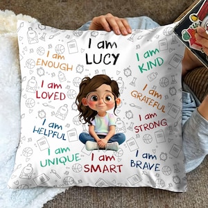 May include: A white cushion with a cartoon illustration of a girl wearing glasses. The cushion has the text "I am LUCY" and other positive affirmations such as "I am KIND" and "I am BRAVE". The background is decorated with small doodle illustrations.