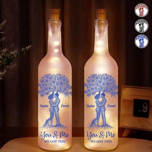 May include: Two illuminated glass bottles with cork stoppers. Each bottle features a blue tree design with a couple embracing, the names "Sophia" and "Daniel", and the text "You & Me WE GOT THIS". The bottles are lit from within.