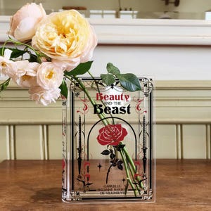 K&ouml;nnte beinhalten: Eine durchsichtige Glasvase in Form eines Buches mit dem Titel "Die Sch&ouml;ne und das Biest" auf der Vorderseite. Die Vase ist mit einem Strau&szlig; blassgelber Rosen und einer einzelnen roten Rose gef&uuml;llt.