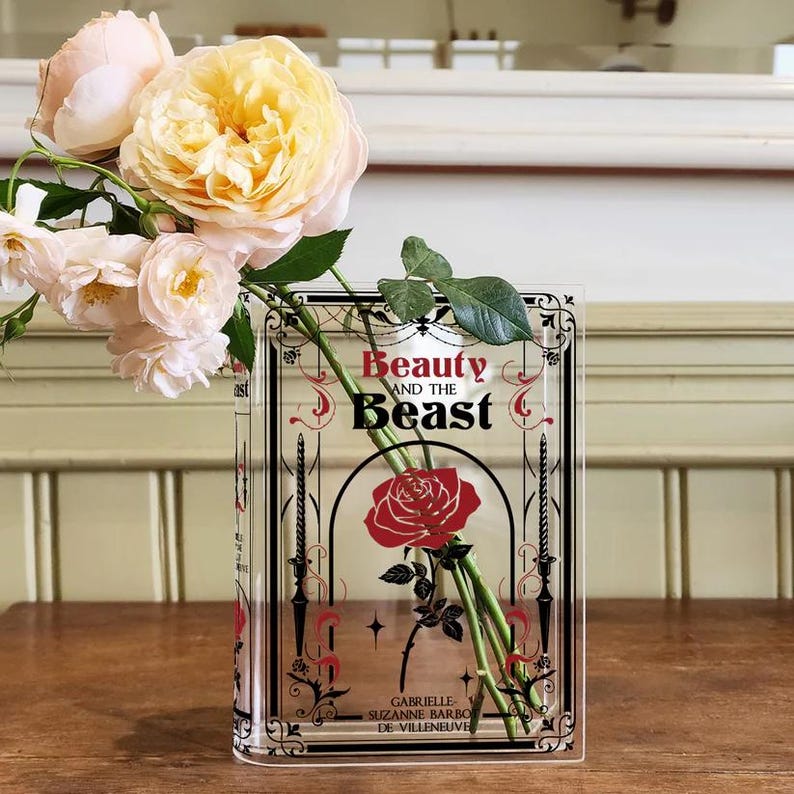 K&ouml;nnte beinhalten: Eine durchsichtige Glasvase in Form eines Buches mit dem Titel "Die Sch&ouml;ne und das Biest" auf der Vorderseite. Die Vase ist mit einem Strau&szlig; blassgelber Rosen und einer einzelnen roten Rose gef&uuml;llt.