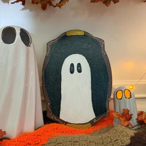 Könnte beinhalten: Eine Halloween-Dekoration mit drei Geisterfiguren. Das Herzstück ist ein gerahmtes Geistergemälde mit einem weißen Geist auf dunklem Hintergrund. Zwei Keramik-Geisterfiguren, eine große und eine kleine, stehen auf einer bunten Decke mit Herbstblättern.