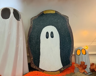 Può includere: Un'esposizione a tema Halloween con tre figure di fantasmi. Il pezzo centrale è un dipinto di fantasmi incorniciato con un fantasma bianco su sfondo scuro. Ai lati ci sono due figure di fantasmi in ceramica, una grande e una piccola, su una coperta colorata con foglie autunnali.