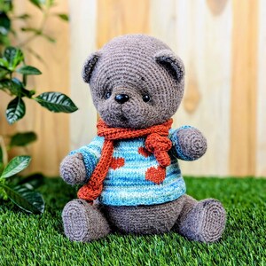 May include: A gray crocheted teddy bear wearing a blue and white striped sweater with red polka dots and a red scarf. The bear is sitting on green grass.
