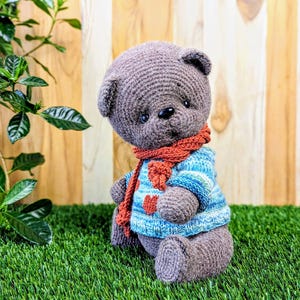 May include: A gray crocheted teddy bear wearing a blue and white striped sweater with a red scarf. The bear is sitting on a green grassy surface.