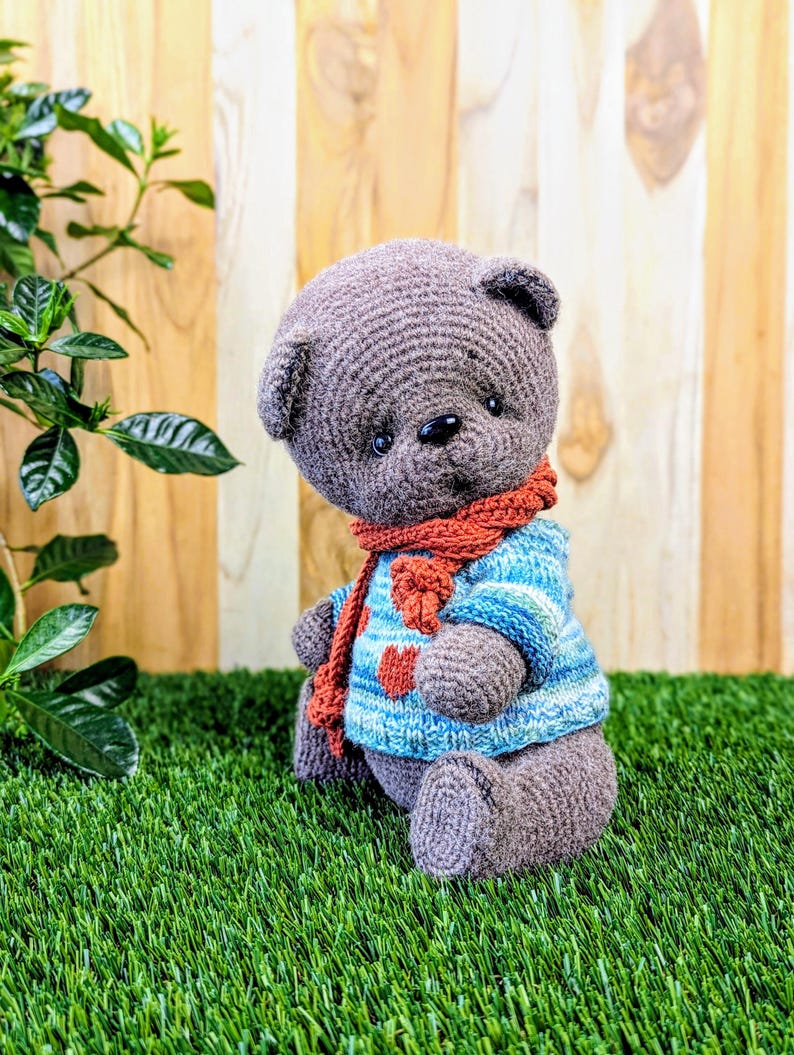 May include: A gray crocheted teddy bear wearing a blue and white striped sweater with a red scarf. The bear is sitting on a green grassy surface.