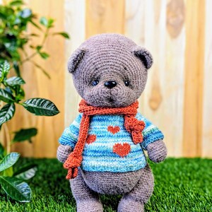 May include: A gray crocheted teddy bear wearing a blue and white striped sweater with red hearts and a red scarf. The bear is standing on green grass.