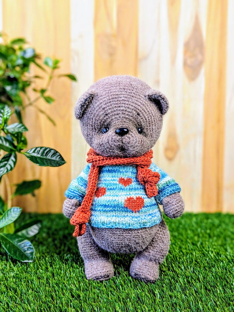 May include: A gray crocheted teddy bear wearing a blue and white striped sweater with red hearts and a red scarf. The bear is standing on green grass.