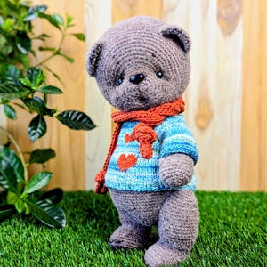 May include: A crocheted gray teddy bear wearing a blue and white striped sweater with a red heart design and a red scarf. The bear is standing on green grass.