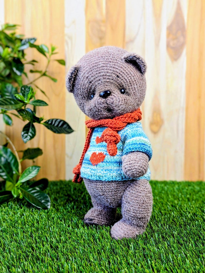 May include: A crocheted gray teddy bear wearing a blue and white striped sweater with a red heart design and a red scarf. The bear is standing on green grass.