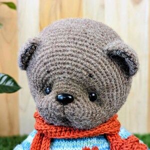 May include: A brown crocheted teddy bear wearing a blue and white striped sweater with red hearts and a red scarf.