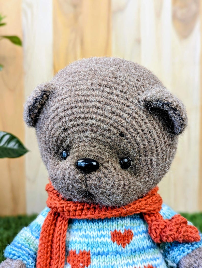 May include: A brown crocheted teddy bear wearing a blue and white striped sweater with red hearts and a red scarf.