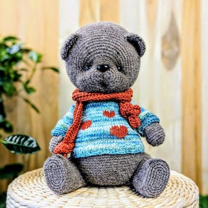 May include: A gray crocheted teddy bear wearing a blue and white striped sweater with red hearts and a red scarf. The bear is sitting on a woven wicker stool.