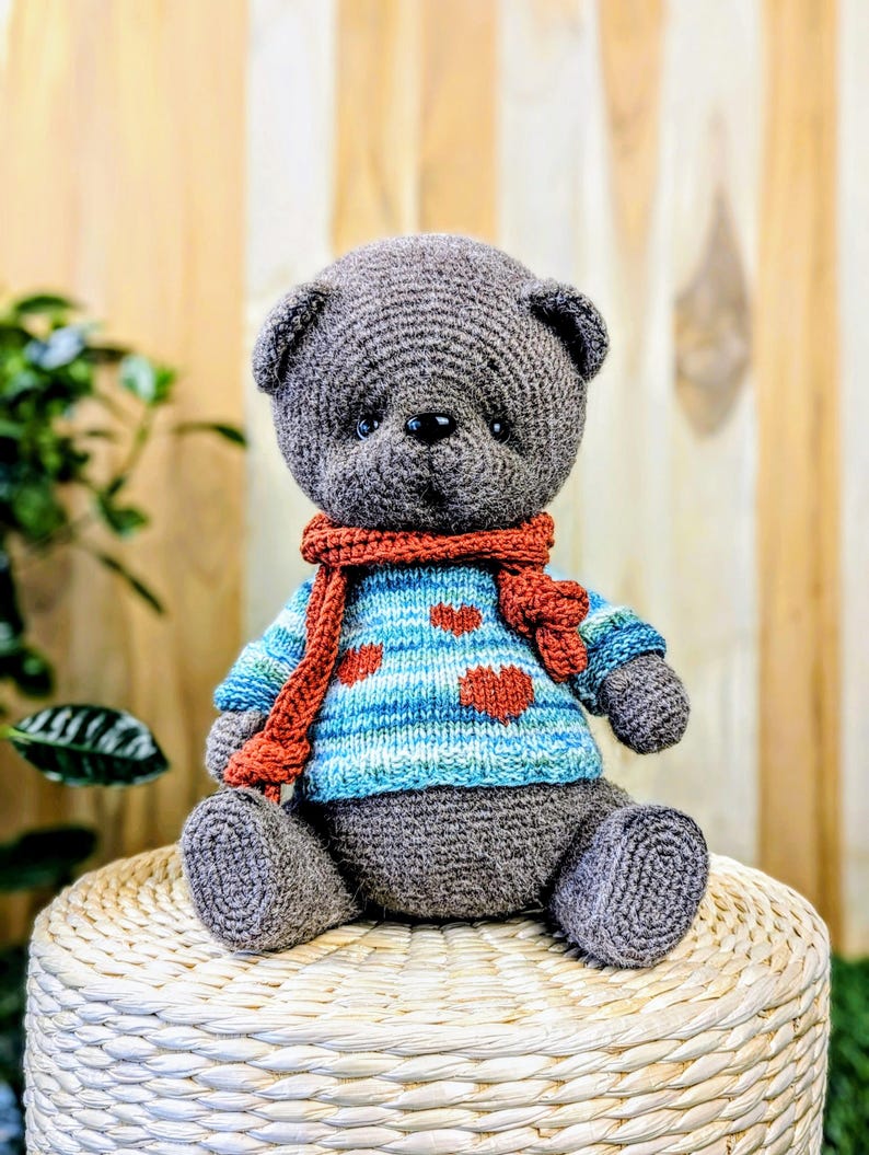 May include: A gray crocheted teddy bear wearing a blue and white striped sweater with red hearts and a red scarf. The bear is sitting on a woven wicker stool.