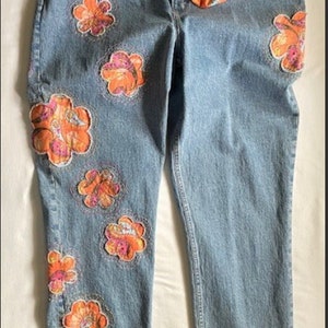 May include: Light blue denim jeans adorned with orange and pink floral appliqués. A matching scrunchie rests on the jeans. The jeans have a classic five-pocket design and a relaxed fit, perfect for casual wear.