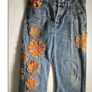 以下が含まれることがあります： 左足にカラフルな花の刺繍が施された青いデニムのジーンズ。刺繍はオレンジ、イエロー、レッド、グリーンの色合いです。