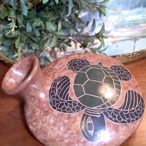 May include: A decorative, rounded vase with a turtle design. The vase is a mottled reddish-brown color with a dark green turtle design. The turtle's shell is segmented, and its flippers are outlined in black. The vase has a curved spout.