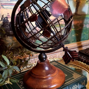 May include: A metal globe with a wooden base and an arrow pointing to the right. The globe is made of wire and has a red, brown, and gold color scheme. The globe is sitting on top of two green books with gold accents.