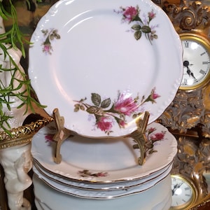 May include: A stack of white porcelain plates with a scalloped edge and a floral rose design in pink and green. The plates are displayed on a gold-colored metal stand. A vintage clock and decorative tray are in the background.