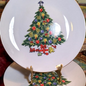 May include: Two white ceramic plates with gold rims, each featuring a Christmas tree design. The tree is decorated with ornaments, candles, and a toy train. A small angel tops the tree. The plates are displayed on a gold-colored stand.