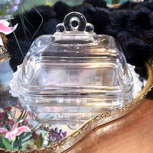 May include: A clear, square glass butter dish with a lid, featuring a loop handle and decorative accents. The dish sits on a gold-colored tray with floral accents and a white lace trim. The background includes a painting and a black fur-like material.