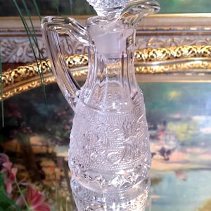 May include: A clear glass bottle with a textured surface and a decorative stopper. The bottle has a handle and a round base. The bottle is sitting on a gold-colored surface.