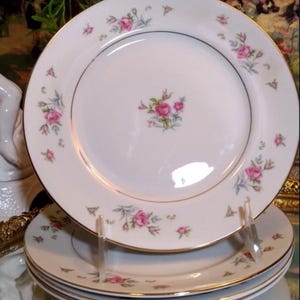 May include: Set of four white porcelain dinner plates with a delicate floral design featuring pink roses and green leaves. The plates have a gold trim around the edge.