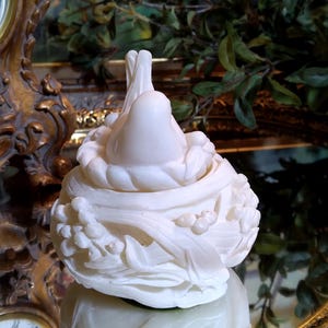 May include: A white, decorative sculpture of a bird in a nest. The sculpture is made of a smooth, possibly ceramic material, and features intricate details of leaves and berries. The sculpture is reflected in a mirrored surface.