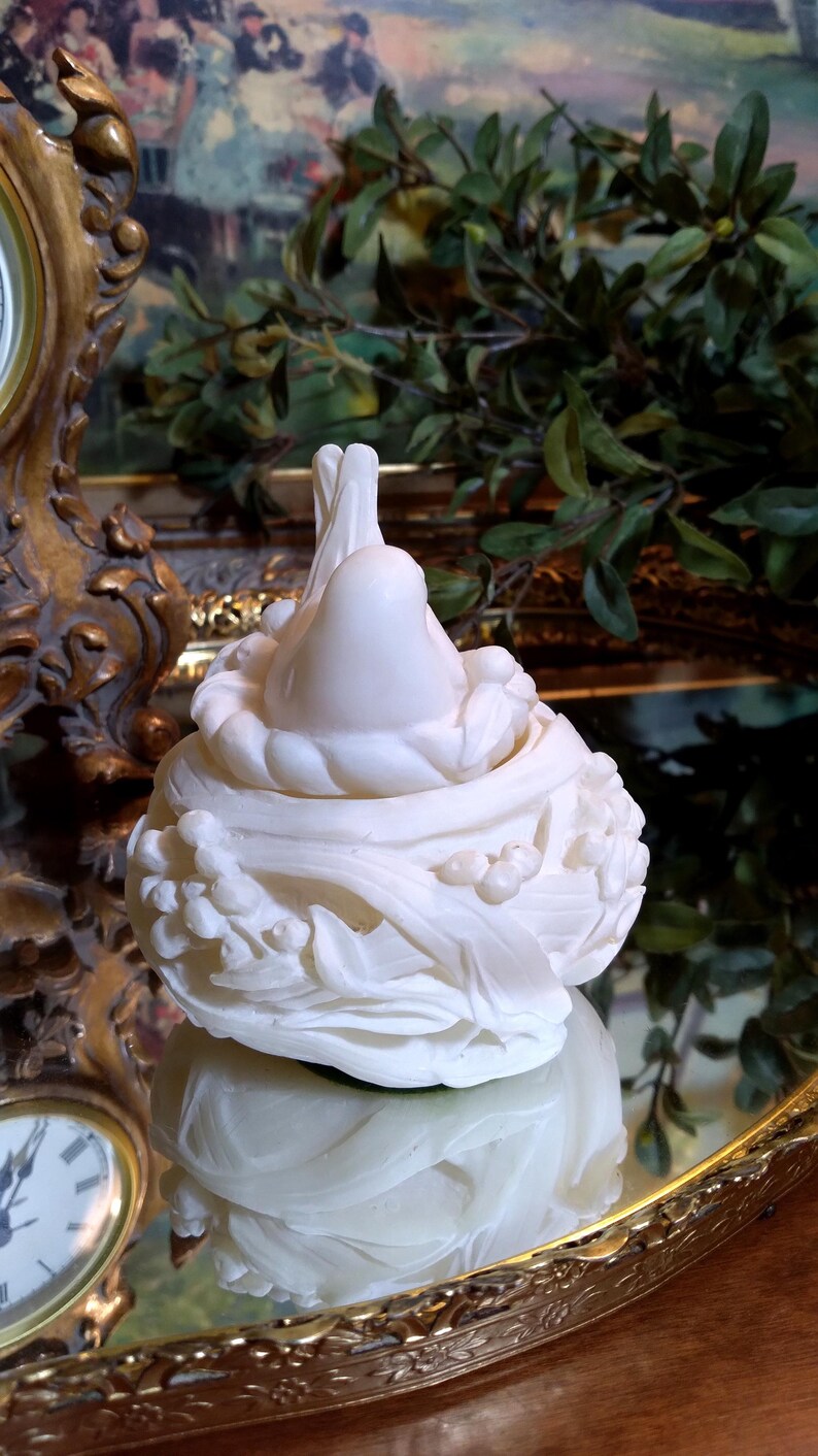 May include: A white, decorative sculpture of a bird in a nest. The sculpture is made of a smooth, possibly ceramic material, and features intricate details of leaves and berries. The sculpture is reflected in a mirrored surface.