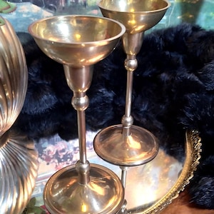 May include: Two vintage brass candle holders with a classic design. The candle holders have a round base, a slender stem, and a wide, shallow cup to hold the candle. The candle holders are displayed on a decorative tray.