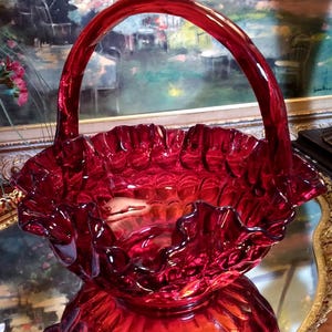 May include: A ruby red glass basket with a ruffled rim and a handle. The basket is sitting on a gold-colored tray.