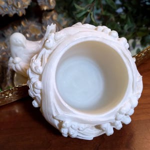 May include: Ivory-colored, ornate decorative container with a circular opening. The container features detailed relief carvings of floral and foliate designs. The background includes a gold-framed clock and greenery.