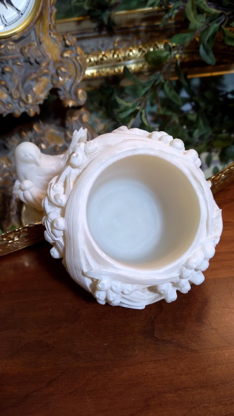 May include: Ivory-colored, ornate decorative container with a circular opening. The container features detailed relief carvings of floral and foliate designs. The background includes a gold-framed clock and greenery.