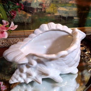 May include: A white ceramic seashell planter with a smooth, glossy finish. The planter is shaped like a large seashell with a wide opening at the top.