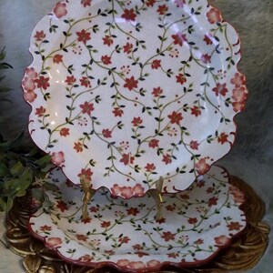 May include: Two white ceramic plates with scalloped edges, featuring a repeating pattern of small red flowers and green vines. The plates are displayed on a gold-toned stand, with a red painted trim along the scalloped rim of each plate.