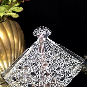 Peut inclure: Un plat en verre transparent en forme d'éventail avec un motif en étoile détaillé. Le plat a une poignée décorative et repose sur une base marron. L'arrière-plan comprend un vase doré avec des fleurs rouges.