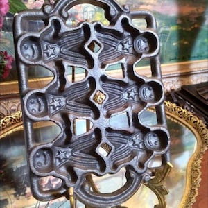 May include: A cast iron mold for making gingerbread men. The mold is in the shape of six gingerbread men, each with a smiling face, a star on their chest, and a button on their belly. The mold is on a gold stand.