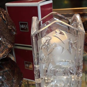 May include: Clear glass candle holder with etched holiday scenes. The candle holder has a rectangular shape with a roof-like top. A red box with the Mikasa logo is visible in the background.