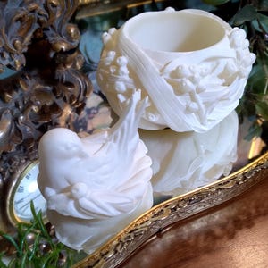 May include: Two white, ornate, decorative objects. One is a small, open-topped bowl with floral relief. The other is a sculpted bird. Both are on a mirrored surface, with a gold-toned, ornate clock and tray in the background.