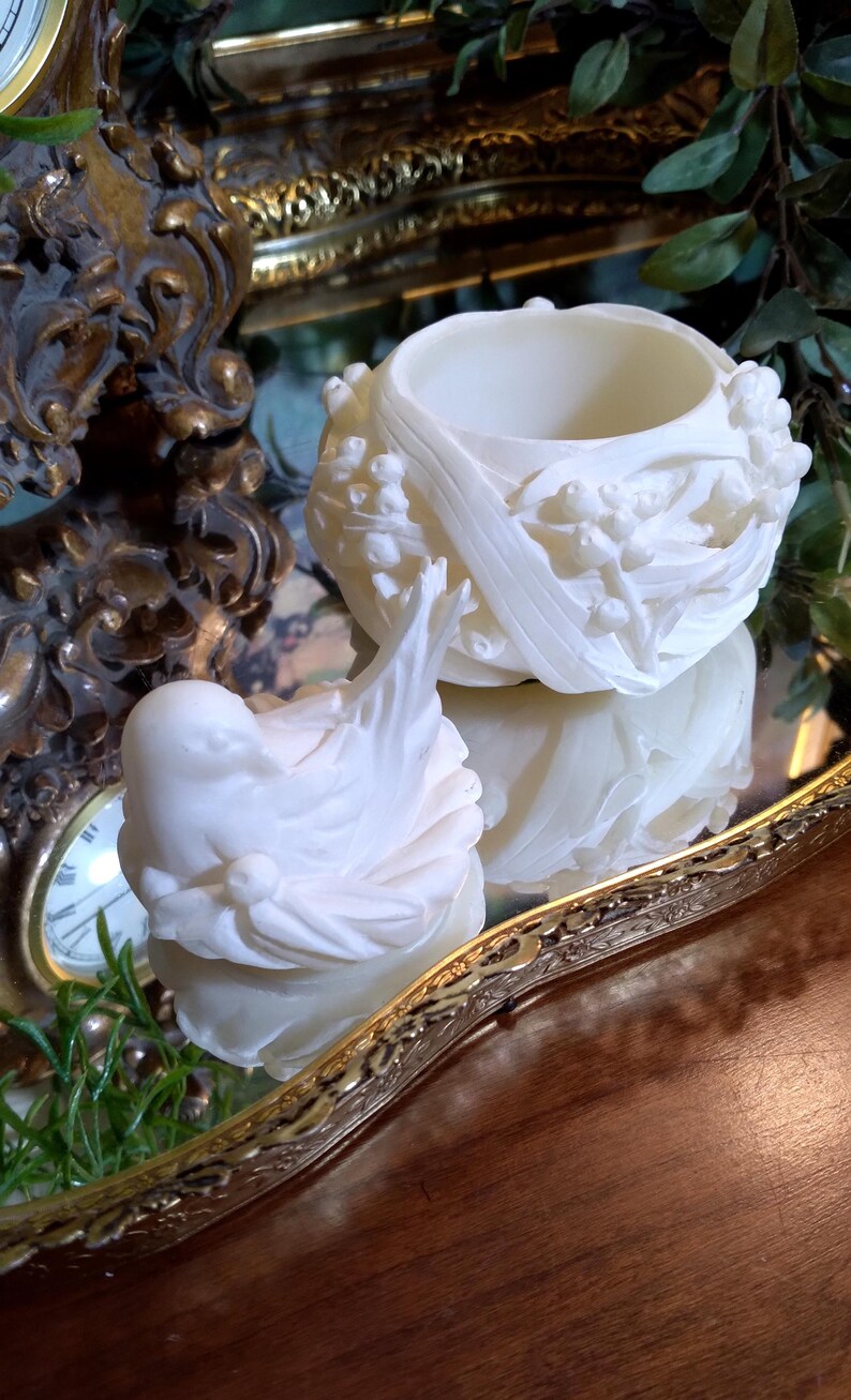 May include: Two white, ornate, decorative objects. One is a small, open-topped bowl with floral relief. The other is a sculpted bird. Both are on a mirrored surface, with a gold-toned, ornate clock and tray in the background.