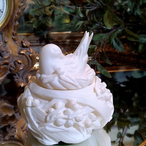 May include: A white, sculpted bird figurine in a nest, with detailed feather and floral designs. The bird is perched atop a nest-like base, creating a decorative accent piece. The figurine is likely made of ceramic or porcelain.