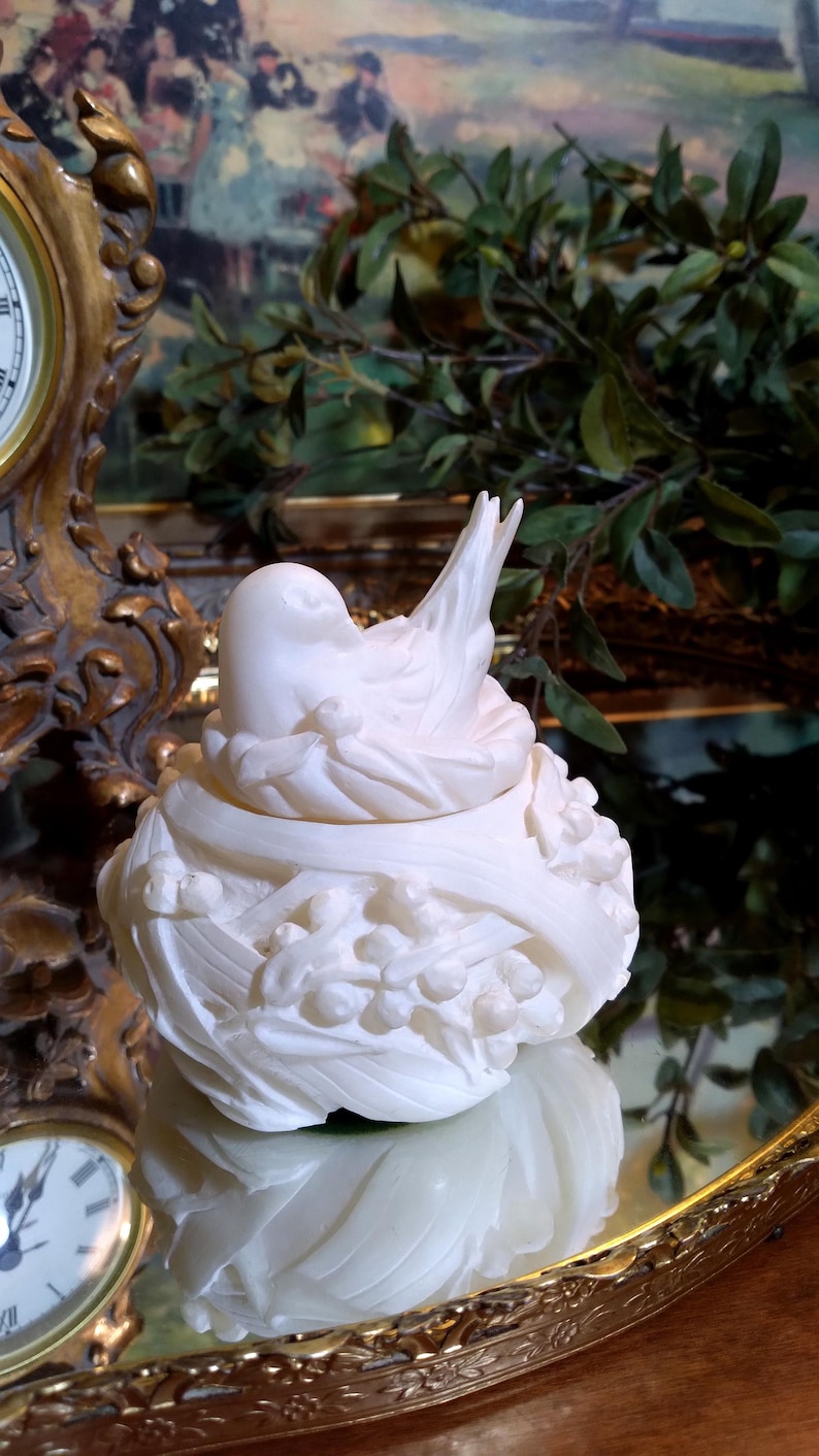 May include: A white, sculpted bird figurine in a nest, with detailed feather and floral designs. The bird is perched atop a nest-like base, creating a decorative accent piece. The figurine is likely made of ceramic or porcelain.
