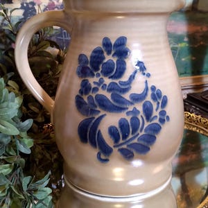 May include: A beige ceramic pitcher with a blue floral design. The pitcher has a curved handle and a slightly flared rim. The blue floral design is centered on the front of the pitcher. The pitcher is sitting on a mirrored surface.