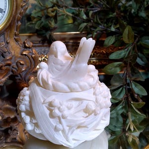 May include: A detailed, off-white bird-shaped trinket box. The lid features a bird perched atop a nest, with floral and leaf detailing around the base. The box is displayed on a reflective surface, with a blurred background.