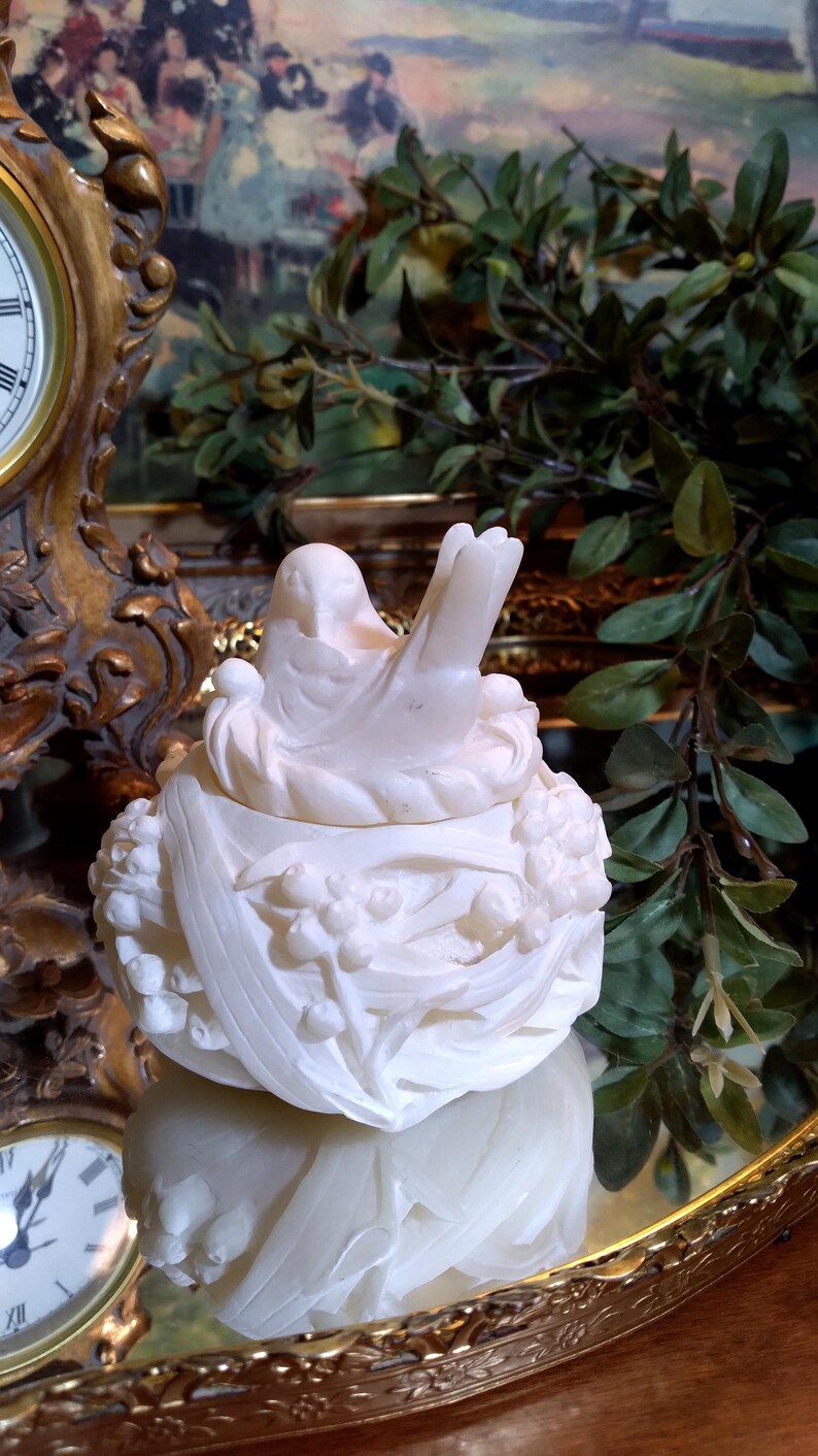 May include: A detailed, off-white bird-shaped trinket box. The lid features a bird perched atop a nest, with floral and leaf detailing around the base. The box is displayed on a reflective surface, with a blurred background.