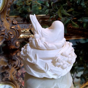May include: A white ceramic bird figurine perched on a nest-shaped base. The bird has a smooth, rounded body and is nestled among detailed floral and leaf accents. The figurine is displayed on a reflective surface, with a gold-framed clock in the background.